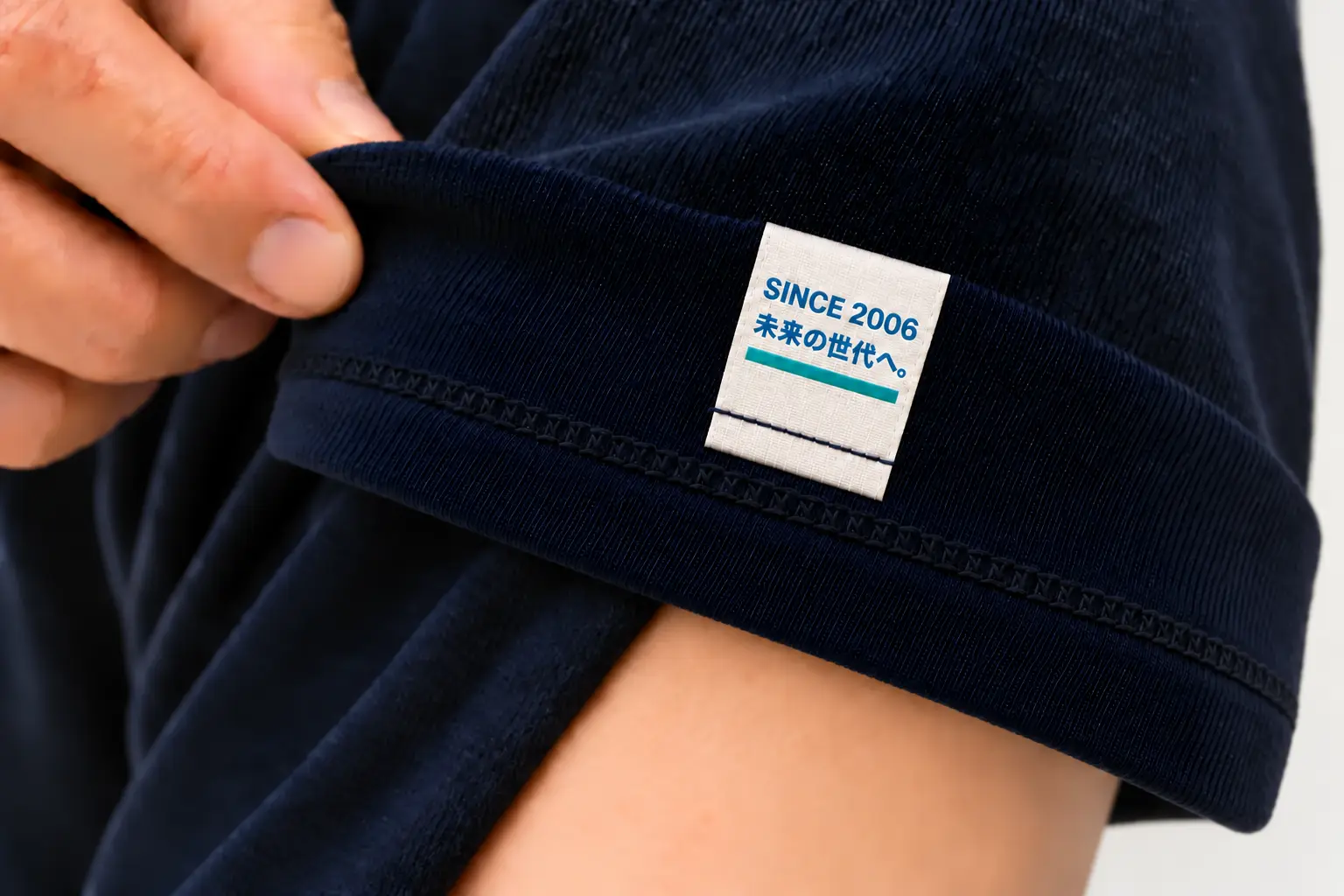濃紺Tシャツの袖口を外側へ折り返し、縦長タグ裏面のSINCE 2006と未来の世代へというメッセージを見せている写真