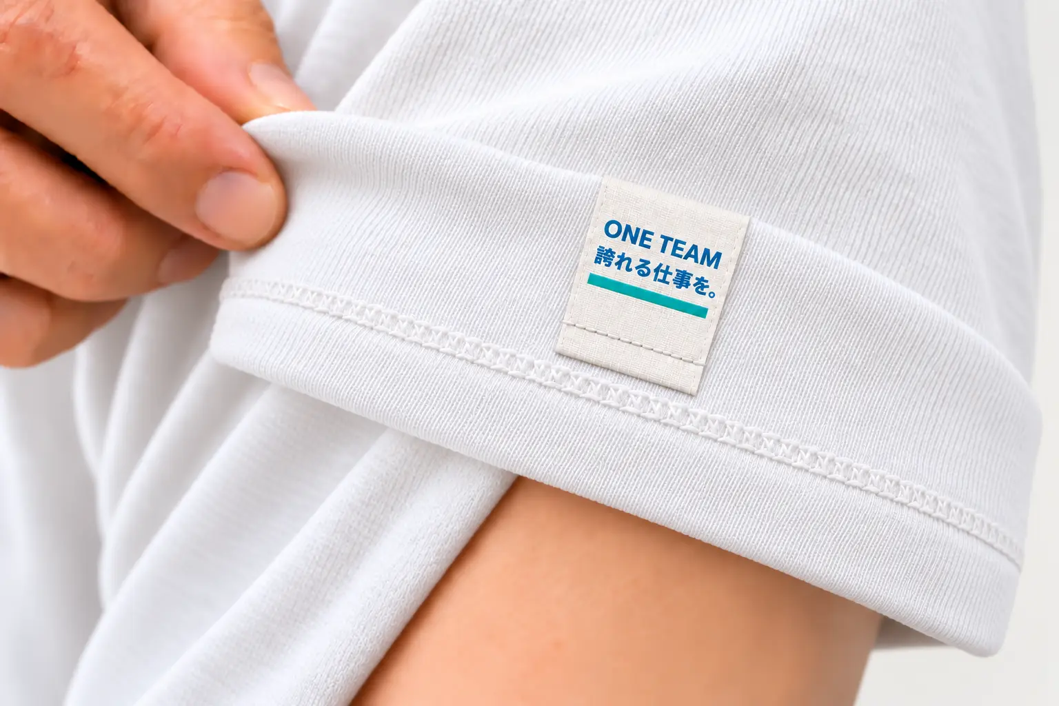 白いTシャツの袖口を外側へ折り返し、縦長タグ裏面のONE TEAMと誇れる仕事をというメッセージを見せている写真